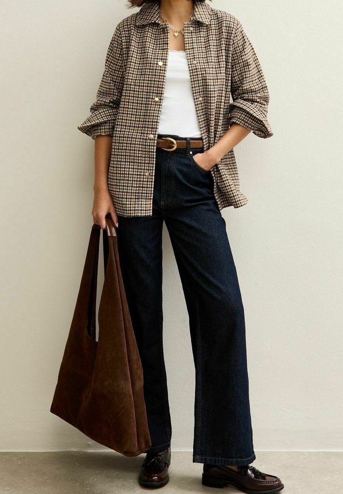 Femme portant une chemise à carreaux boutonnée, un haut blanc, un jean foncé, une ceinture et des chaussures marron, tenant un grand sac fourre-tout marron à ses côtés.