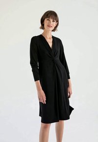 Vestido preto até ao joelho com mangas longas, apresentando um detalhe frontal torcido, tecido suave e um corte descontraído. Textura lisa, design minimalista.