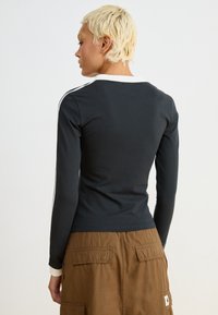 Camisola preta de mangas longas com colarinho branco e riscas laterais. Tecido macio, design ajustado. Combinada com uma saia castanha com detalhes de bolsos.