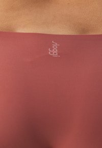 Rotes, figurbetontes Kleidungsstück aus glattem Stoff, mit einem horizontal in Weiß bedruckten Logo in der Nähe der Oberkante. Die Textur wirkt weich und dehnbar.