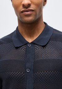 Εγγύς πλάνο ενός άνδρα που φοράει μπλε σκούρο, κουμπωτό, πλεκτό πόλο με διάτρητο ύφασμα και γιακά, με μονόχρωμο φόντο.