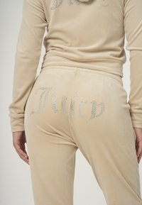Beige bársonyos melegítő, a nadrág hátulján strasszkövekből készült betűzéssel, ami "Juicy" feliratot formáz, puha, sima textúrával.