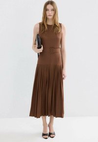 Bruine mouwloze midi-jurk met een geplooide rok, gestructureerd lijfje en een getailleerde taille met ceintuur. Gecombineerd met een zwarte clutch en sandalen met hak.