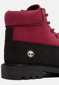 Botín de ante burdeos con acentos de cuero negro, cordones redondos, suela de goma y un logo blanco en el costado. Cuello ajustado y detalles de costura.