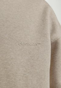 Tan sweatshirt fabric with a soft texture, featuring the word "ORIGINALS™" embossed in a lighter shade on the chest area.