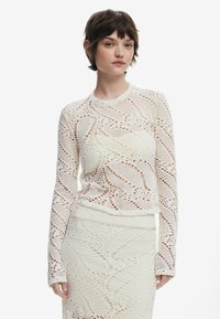 Maglietta bianca a maniche lunghe con un motivo in pizzo lavorato a maglia, caratterizzata da intricati intagli e un colletto rotondo, abbinata a una gonna coordinata.
