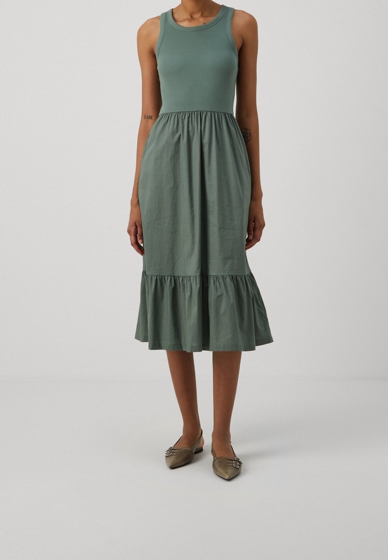 Robe midi verte sans manches avec taille froncée et ourlet volanté portée avec des chaussures plates pointues marron sur une personne debout.