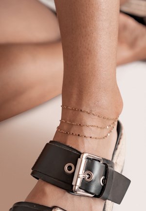 Black leather anklet strap with silver hardware, featuring three thin gold chains around the ankle, highlighting skin texture.