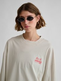 Mlada oseba s kratkimi rjavimi lasmi, ki nosi majhna ovalna črna sončna očala, zlate obroče uhanov in kremno srajco z rdečim napisom na prsih.