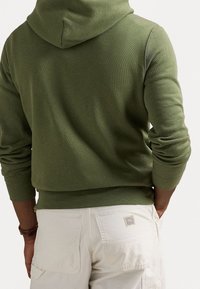 Sudadera verde oliva con capucha en la espalda y puños acanalados, combinada con pantalones de color claro que tienen una etiqueta en el bolsillo trasero. Tejido texturizado.