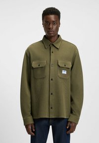 Olive green, ribbed button-up shirt featuring two chest pockets, black buttons, and a small logo patch. Made from textured fabric.