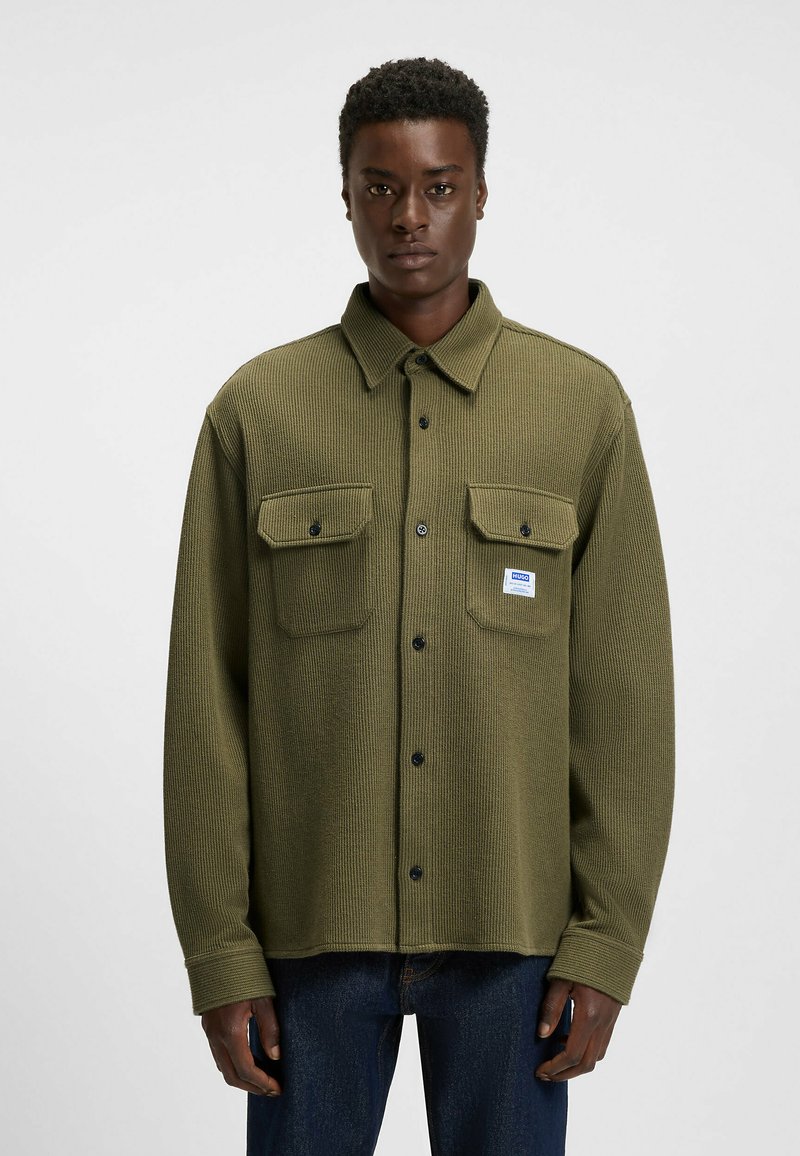 Olive green, ribbed button-up shirt featuring two chest pockets, black buttons, and a small logo patch. Made from textured fabric.