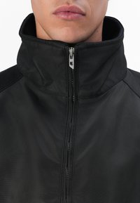 Black zip-up jacket with a high collar, featuring smooth fabric, a front zipper, and subtle stitching details along the seams.