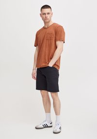 Camisa de tirantes cortos color óxido con detalle de bolsillo, combinada con pantalones cortos oscuros y zapatillas blancas, con acentos en negro y gris y calcetines de caña corta.