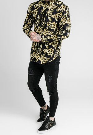 Mężczyzna w czarnej i złotej koszuli z wzorem zapinanej na guziki, podartych czarnych obcisłych dżinsach i czarnych trampkach z białą podeszwą, stojący z zespolonymi dłońmi.