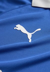 Mėlyna sportinė apranga su tekstūruota, kvėpuojančia medžiaga, ant kurios yra baltas Puma logo. Dizainas apima subtilius tinklinio raštus.