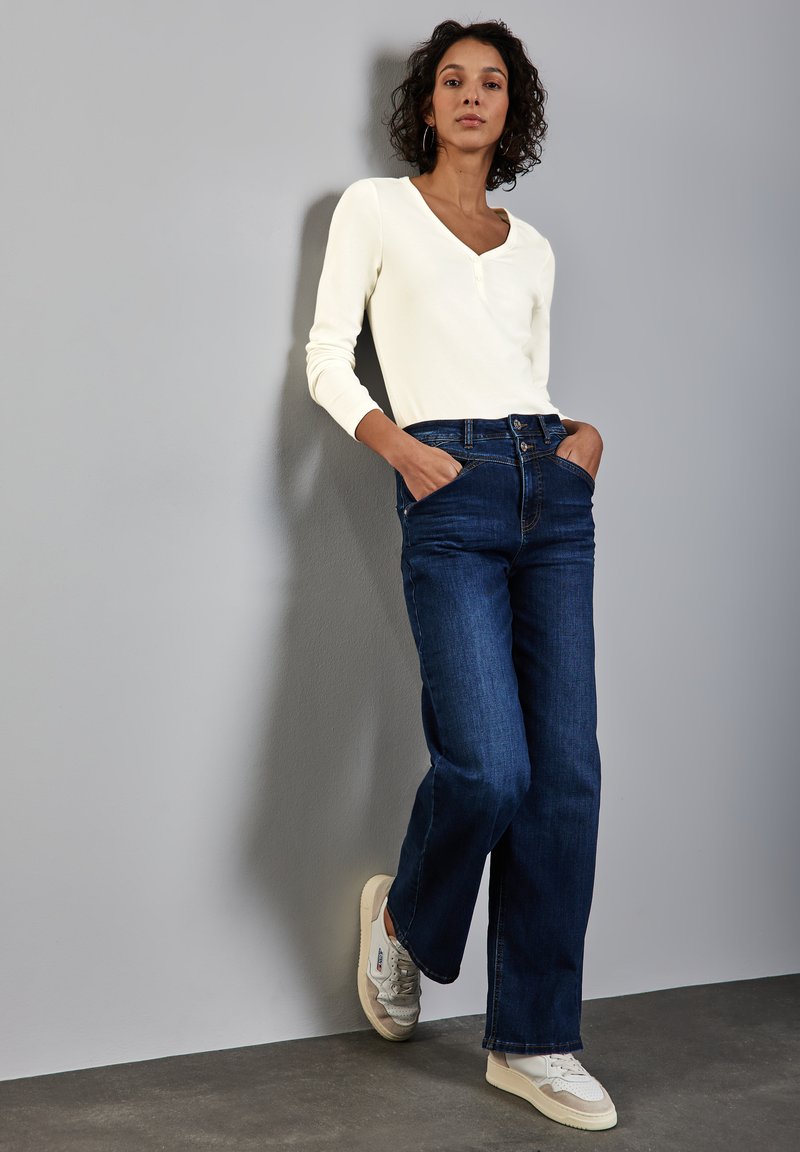 Vit långärmad topp med rund halsringning, kombinerad med mörkblå jeans med raka ben och ljusa sneakers. Mjukt tyg, avslappnad passform.