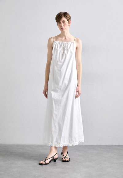 Weißes Baumwoll-Maxi-Kleid mit elastischem Halsausschnitt und Kordelzugdetail. Mit Spitzensaum am Saum verziert. Kombiniert mit schwarzen Sandalen mit Riemchen und Absatz.