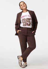 Bruin op maat gemaakt pak met een witte graphic t-shirt met rode en luipaardprintaccenten. Schoeisel is gepatroniseerd in wit en bruin.