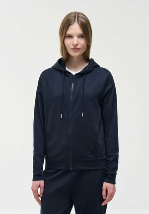 Młoda kobieta z prostymi włosami, ubrana w czarną bluzę z kapturem na zamek i dopasowane czarne spodnie, stojąca na jednolitym jasnym tle.