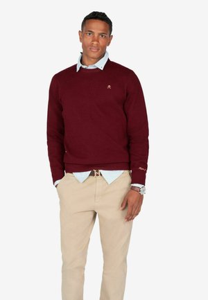 Suéter de punto burdeos con cuello redondo, camisa de color azul claro debajo y pantalones caqui; presenta un sutil detalle del logotipo en el suéter.