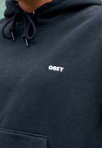 Černá mikina s klokaní kapsou; má bílé vyšívané logo "OBEY" na hrudi a stahovací šňůrky s černými plastovými konci.