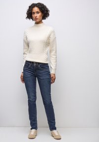 Vit stickad tröja med upphöjt mönster, hög halsringning, kombinerad med mörkblå jeans och beige sneakers. Enskild figur mot en enkel bakgrund.