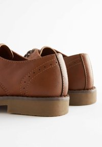 Chaussures en cuir marron avec une texture lisse, un bout rond, des coutures détaillées, et une semelle en caoutchouc basse. Comprend des accents perforés le long des coutures.
