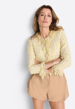 Woman with loose wavy hair wearing a yellow textured fringe jacket and beige high-waisted shorts, arms crossed, standing against a white background.