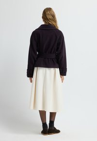 Mörklila ytterplagg med bälte i midjan och breda manschetter, ihop med en flödande vit kjol. Mjuk textur, enkel design, inga synliga mönster.