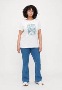 Hvit t-skjorte med et turkist geometrisk mønster på fronten, kombinert med blå utsvingte jeans; casual passform, korte ermer, bomullskvalitet.