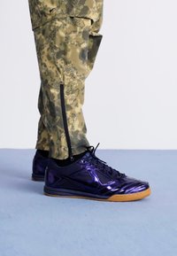 Μωβ αθλητικά παπούτσια με γυαλιστερή επιφάνεια και σόλα από καουτσούκ, σε συνδυασμό με παντελόνι καμουφλάζ με φερμουάρ στα πλάγια.