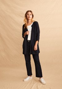 Dunkelblauer Cardigan mit Dreiviertelärmeln, offener Vorderseite, zwei Fronttaschen, getragen über einem weißen T-Shirt und kombiniert mit dunklen Jeans und weißen Sneakers.