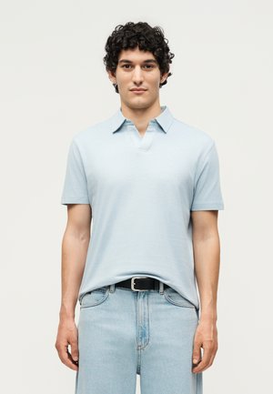 Giovane uomo con capelli ricci e scuri che indossa una polo azzurro chiaro a maniche corte infilata in jeans chiari con cintura nera, in posizione neutra.