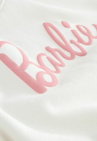 Material alb care prezintă un logo roz „Barbie” reliefat, scris cu un font cursiv, având o textură netedă și o distribuție uniformă a culorii.