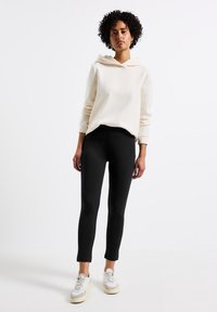 Felpa beige chiaro con scollatura tonda e spalle scese, abbinata a leggings neri aderenti e sneaker bianche con dettagli beige.