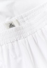 Ceinture élastique blanche avec tissu froncé et une petite étiquette blanche arborant un logo noir brodé.