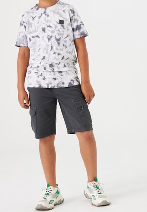 Koszulka młodzieżowa w stylu tie-dye w kolorach białym i szarym, zestawiona z ciemnoszarymi szortami cargo z bocznymi kieszeniami, uzupełniona masywnymi białymi sneakersami.