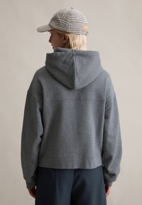 Grijze cropped hoodie met capuchon, met een gladde textuur. Draagt een beige geruit petje. Achteraanzicht, met de nadruk op de casual pasvorm.