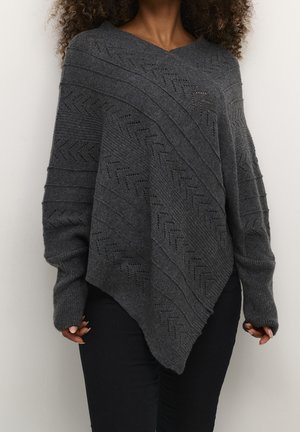 Femme portant un pull poncho en maille gris foncé avec des motifs texturés en diagonale et des manches longues, associé à un pantalon noir.