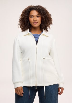 Frau mit lockigem Haar, die eine weiße Jacke mit Reißverschluss und Kordelzug sowie blaue Jeans trägt, steht vor einem schlichten hellen Hintergrund.