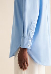Camicia azzurra a maniche lunghe con orlo curvato e polsini abbottonati, realizzata in un tessuto liscio. Presenta una vestibilità rilassata e dettagli minimi.