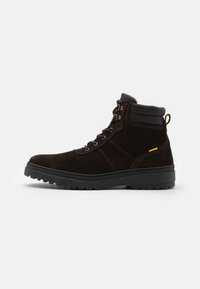 Brown suede ankle boot with a black rubber sole, metal eyelets, padded collar, and a small yellow accent on the side. Flat laces.