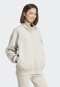 Svetlo béžová zipová mikina s vysokým golierom, vo voľnom strihu a so zúženými manžetami. Mäkký materiál; má bočné vrecká a hladkú textúru.