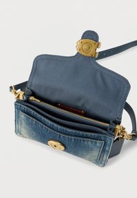 Majhna modra denim torbica čez ramo z zlatim zaponko, odprt zgornji del, ki razkriva notranji žep z zadrgo in usnjen pas na belem ozadju.
