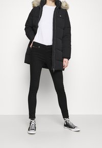 Osoba v černé péřové bundě s kožešinovou kapucí, bílem tričku, černých skinny kalhotách a černých vysokých teniskách s bílými tkaničkami.