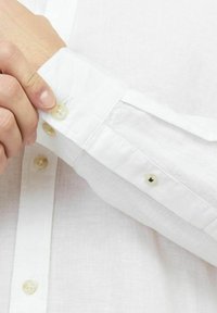 Camicia di lino bianca con polsino a tre bottoni, cuciture visibili e superficie in tessuto testurizzato per una buona traspirabilità.