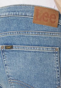 Jeans de mezclilla de color azul claro con un parche de cuero etiquetado como "Lee" en la cinturilla y una pequeña etiqueta negra en el bolsillo trasero.