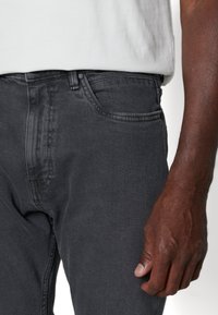 Jeans de mezclilla de color gris oscuro con un diseño estándar de cinco bolsillos y una superficie texturizada, mostrados con una mano descansando en la zona del muslo.