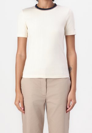 Osoba ubrana w kremowy, prążkowany top z krótkim rękawem, z granatowo-czerwonym wykończeniem kołnierza, w połączeniu z beżowymi spodniami, ramiona swobodnie opuszczone wzdłuż ciała.